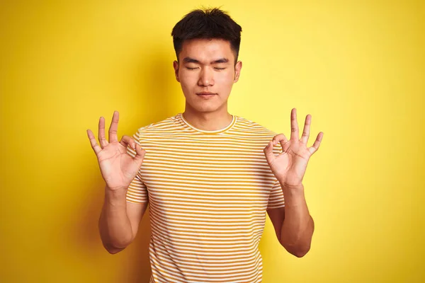 孤立した黄色の背景の上に立つTシャツを着た若いアジアの中国人男性は 指で瞑想のジェスチャーをして目を閉じて微笑んでいます ヨガのコンセプト — ストック写真
