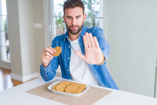 Handsome man eating healthy whole grain biscuit with open hand doing stop sign with serious and confident expression, defense gesture
