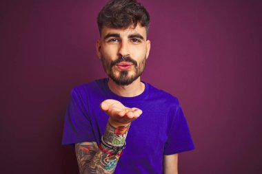 Dövme giyen genç adam izole mor arka plan üzerinde duran t-shirt hava güzel ve seksi olmak eliyle bir öpücük üfleme kameraya bakarak. Aşk ifadesi.