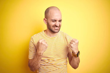 Sakallı genç kel adam sarı izole arka plan üzerinde rahat çizgili t-shirt giyen çok mutlu ve heyecanlı kolları kaldırdı ile kazanan jest yapıyor, gülümseyerek ve başarı için çığlık. Kutlama konsepti.