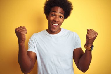 Afro saçlı beyaz t-shirt giyen Amerikalı adam izole sarı arka plan üzerinde ayakta sürpriz ve kollarını kaldırdı ve açık gözleri ile başarı için şaşırttı. Kazanan kavramı.