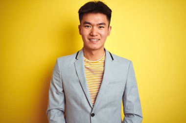 Genç Asyalı Çinli işadamı yüzünde mutlu ve serin bir gülümseme ile izole sarı arka plan üzerinde duran ceket giyiyor. Şanslı kişi.