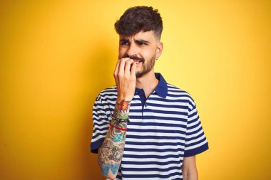İzole sarı arka planda görünümlü çizgili polo ayakta giyen dövme ile genç adam vurguladı ve ağız ısırma çivi elleri ile gergin. Anksiyete sorunu.
