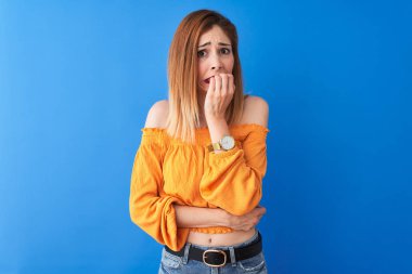Turuncu rahat t-shirt giyen güzel kızıl kadın izole mavi arka plan üzerinde duran stresli ve ağız tırnak ısırma elleri ile sinir görünümlü. Anksiyete sorunu.