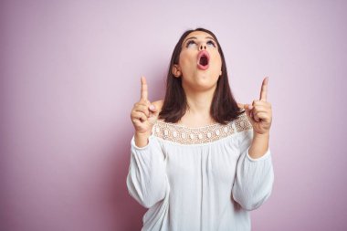 Pembe izole arka plan üzerinde Genç güzel esmer kadın şaşırttı ve yukarı bakarak ve parmakları ile işaret ve kolları kaldırdı şaşırttı.