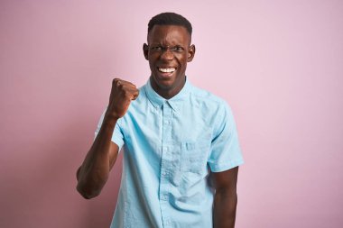 Izole pembe arka plan üzerinde duran mavi rahat gömlek giyen Afro-Amerikan adam kızgın ve öfkeli yükselterek yumruk sinirli ve öfkeli öfke ile bağırırken. Öfke ve agresif kavram.