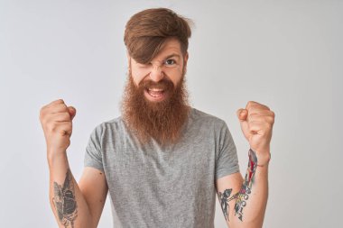 Genç kızıl saçlı İrlandalı adam izole gri arka plan üzerinde duran t-shirt giyen sürpriz ve kollarını kaldırdı ve açık gözleri ile başarı için şaşırttı. Kazanan kavramı.