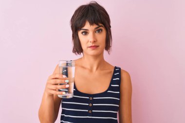 Genç güzel kadın ciddi düşünme akıllı yüz üzerinde emin bir ifade ile izole pembe arka plan üzerinde duran su cam tutan