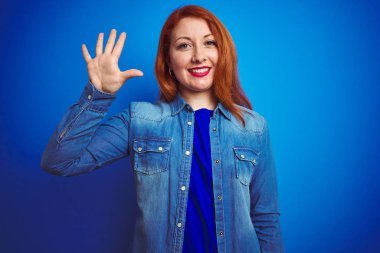Denim gömlek giyen genç güzel kızıl saçlı kadın mavi izole arka plan üzerinde duran gösteren ve emin ve mutlu gülümserken parmakları ile beş işaret.