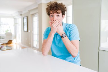 Evde rahat mavi t-shirt giyen genç yakışıklı adam yanlışlık için eller ile ağız kapsayan şok. Gizli konsept.