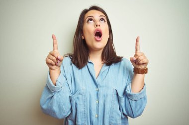Genç güzel esmer kadın izole arka plan üzerinde rahat mavi kot gömlek giyen şaşırmış ve şaşırttı yukarı bakarak ve parmakları ile işaret ve kolları kaldırdı.