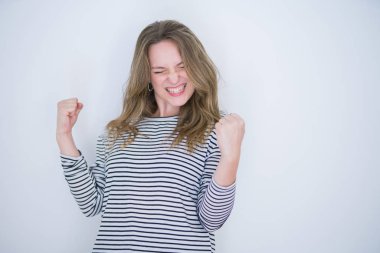 Beautiful blonde girl with blue eyes wearing striped sweater over white isolated background very happy and excited doing winner gesture with arms raised, smiling and screaming for success. Celebration concept.
