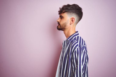 Yan bakarak izole pembe arka plan üzerinde duran çizgili gömlek giyen dövme giyen genç adam, kendine güvenen gülümseme ile doğal yüz ile profil poz dinlenmek.