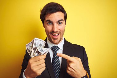 İzole sarı arka plan üzerinde dolar tutan takım elbise giyen genç yakışıklı işadamı el ve parmak ile işaret çok mutlu