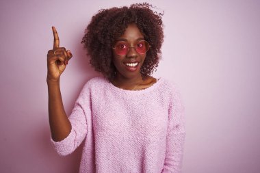 Genç afrikalı afro kadın başarılı bir fikir ile parmak işaret izole pembe arka plan üzerinde kazak ve güneş gözlüğü giyiyor. Çıktı ve mutlu. Bir numara.