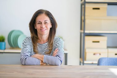 Orta yaş yaşlı kadın evde masada oturan mutlu yüz kameraya bakarak çapraz kolları ile gülümseyerek. Pozitif kişi.