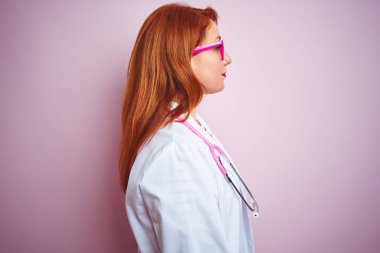 Genç redhead doktor kadın yan bakarak izole pembe arka plan üzerinde duran stetoskop kullanarak, kendine güvenen gülümseme ile doğal yüz ile profil poz dinlenmek.