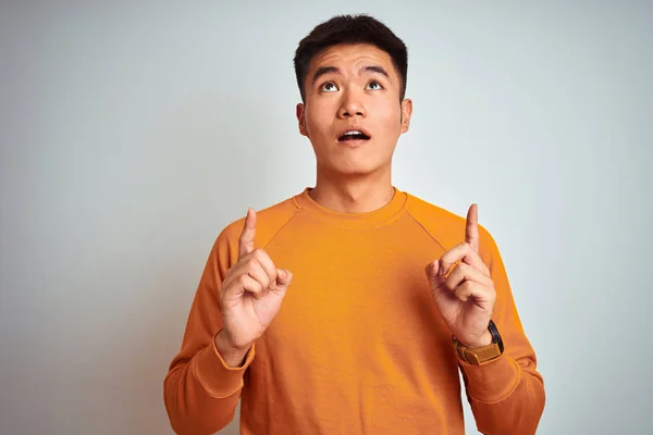 孤立した白い背景の上に立つオレンジ色のセーターを着たアジアの若い中国人男性は 指で見上げて指を指差し 腕を上げて驚いた — ストック写真