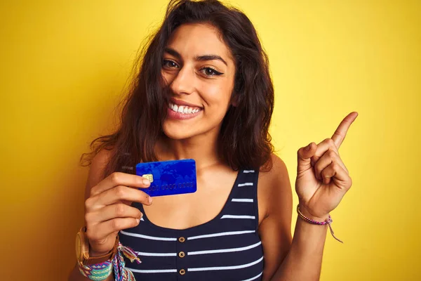 Young beautiful woman holding credit card standing over isolated yellow background very happy pointing with hand and finger to the side