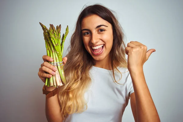 若い美しい女性は 灰色の孤立した背景の上にアスパラガスを食べて 幸せな顔を笑顔で側面に親指で示しています — ストック写真