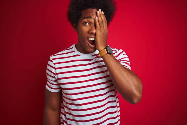 Young african american man with afro hair wearing striped t-shirt over isolated red background covering one eye with hand, confident smile on face and surprise emotion.