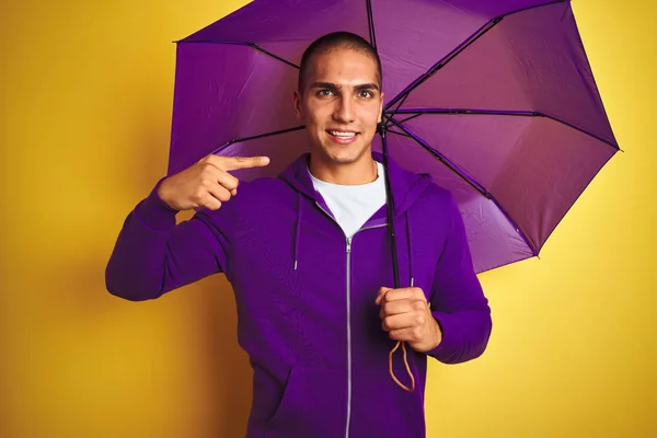 黄色の孤立した背景の上に紫色の傘を身に着けている若いハンサムな男は 手と指で非常に幸せな指差し — ストック写真