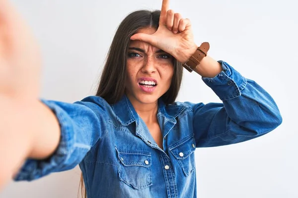 Beautiful woman wearing denim shirt make selfie by camera over isolated white background making fun of people with fingers on forehead doing loser gesture mocking and insulting.