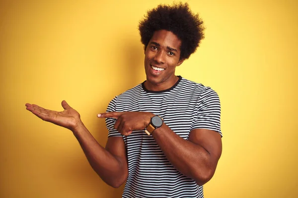 African american man with afro hair wearing navy striped t-shirt over isolated yellow background amazed and smiling to the camera while presenting with hand and pointing with finger.