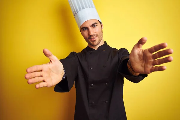 身穿制服和帽子的年轻厨师站在孤立的黄色背景上 看着镜头微笑着张开双臂拥抱 拥抱幸福的欢快表达 — 图库照片