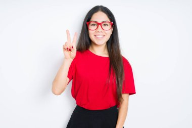 İzole arka plan üzerinde kırmızı gözlük takan güzel esmer kadın zafer işareti yaparak kameraya göz kırparak mutlu yüz ile gülümseyerek. İki numara.