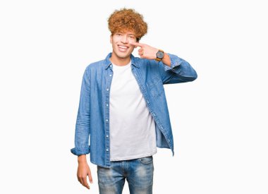 Afro saçlı kot giyen genç yakışıklı adam ceket işaretçi el parmak yüz ve burun, neşeli gülümseyen