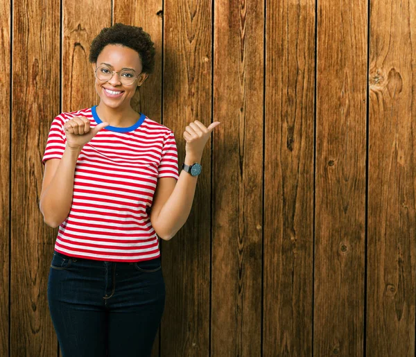 Güzel genç Afro-Amerikan kadın üzerinde gözlük takan izole arka plan emin gülümseyen arkasından el ve kadar başparmak ile işaret