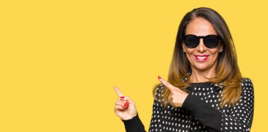 Güzel orta yaş kadın gülümseyerek ve iki elleri ve parmakları ile tarafına işaret kamera bakarak güneş gözlüğü takıyor.