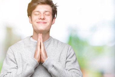 Ellerle birlikte AF emin gülümseyen için soran dua izole arka plan üzerinde sweatshirt giyen genç yakışıklı sportif adam.