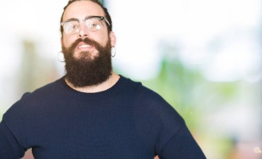 Uzun saç ve sakal uzak tarafına yüzünde, doğal ifade gülümseme ile seyir gözlüklü genç hipster adam. Kendine güvenen gülüyor.