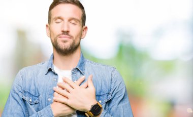 Mavi gözlü ve sakal giyen Denim ceketi olan yakışıklı adam, yüzünde kapalı gözler ve minnettar jest ile göğsünde eller ile gülümsüyor. Sağlık konsepti.