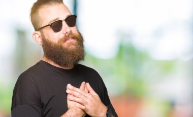 Genç hipster adam gözleri kapalı gözler ve yüz minnettar jest ile göğsünde eller ile gülümseyen güneş gözlüğü giyiyor. Sağlık konsepti.