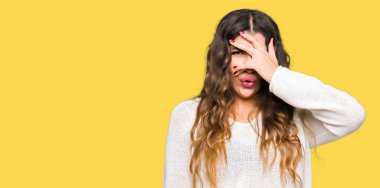 Mahcup ifadeyle parmaklarımın arasından seyir yüz ve gözler el ile kapsayan şok bakmak beyaz kazak giyen genç güzel kadın.