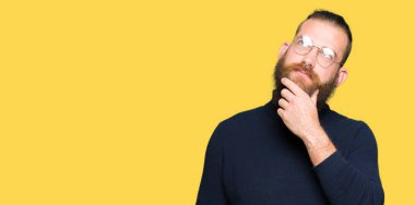 Genç Sarışın giyen gözlük ve balıkçı yaka kazak çene düşünme hakkında soru, dalgın ifade üzerine el ile adam. Düşünceli yüzüyle gülümsüyor. Şüphe kavramı.