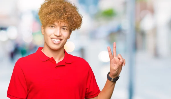 赤いTシャツを着たアフロヘアの若いハンサムな男は 勝利のサインをしているカメラでウインクして幸せそうな顔で微笑んでいます ナンバー2 — ストック写真