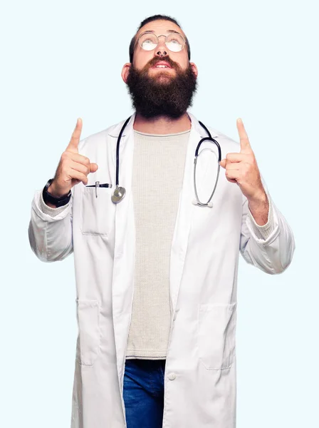 医療のコートを着てひげと若いブロンド医師男びっくりし 驚き見て 指で指して 腕を上げた — ストック写真
