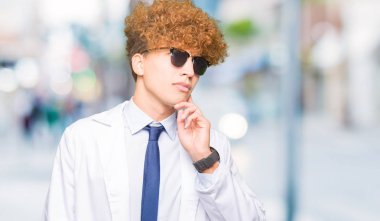 Genç yakışıklı bilim adamı adam profesyonel ceket ve güneş gözlüğü ile el çene üzerinde soru hakkında düşünme, dalgın ifade giyiyor. Düşünceli bir yüz ile gülümsüyor. Şüphe kavramı.