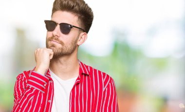 Genç yakışıklı adam çene düşünme hakkında soru, dalgın ifade üzerine el ile güneş gözlüğü takıyor. Düşünceli yüzüyle gülümsüyor. Şüphe kavramı.