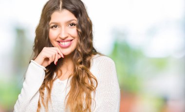 Çene düşünme hakkında soru, dalgın ifade üzerine el ile beyaz kazak giyen genç güzel kadın. Düşünceli yüzüyle gülümsüyor. Şüphe kavramı.