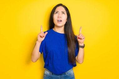 Güzel esmer kadın sarı izole arka plan üzerinde mavi t-shirt giyen şaşırmış ve şaşırmış yukarı bakarak ve parmakları ile işaret ve kolları kaldırdı.