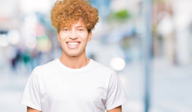 Afro saçlı Genç yakışıklı adam yüz üzerinde mutlu ve serin bir gülümseme ile Casual beyaz t-shirt giyiyor. Şanslı kişi.