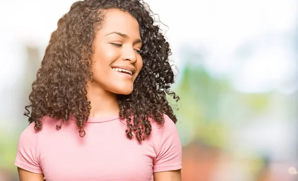 Young beautiful woman with curly hair wearing pink t-shirt looking away to side with smile on face, natural expression. Laughing confident.