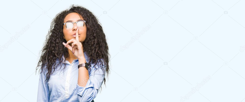 Young beautiful business girl with curly hair wearing glasses asking to be quiet with finger on lips. Silence and secret concept.