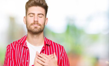 Kırmızı gömlek ile gülümseyen genç yakışıklı adam kapalı gözler ve yüzünde minnettar jest ile göğüs üzerinde eller. Sağlık kavramı.