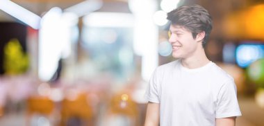 Uzak tarafına yüzünde, doğal ifade gülümseme ile seyir izole arka plan üzerinde rahat beyaz t-shirt giyen genç yakışıklı adam. Kendine güvenen gülüyor.